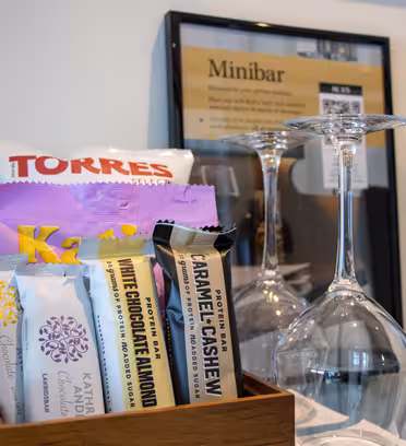 Minibar sortiment med forskellige snacks som chips og chokoladebarer, vinflaske med glas på hovedet og vist minibarmenu i baggrunden.