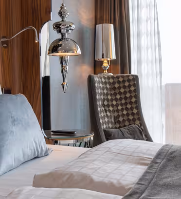 Et moderne hotelværelse med en pæn opredt seng med gråt og hvidt sengetøj og en lyseblå pude. En unik lampe og polstret stol sidder ved siden af et vindue og giver udsigt til murstensbygninger udenfor.