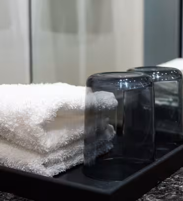 En pænt arrangeret sort bakke på en granitbordplade rummer to foldede hvide håndklæder og to mørke glaskopper på hovedet, med en refleksion synlig i spejlet bag dem.
