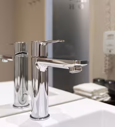 En moderne badeværelsesvask med et elegant krom armatur er vist. Baggrunden har to vægmonterede sæbedispensere, pænt foldede hvide håndklæder og en glasvæg.