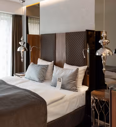 Et moderne hotelværelse med en pænt lavet dobbeltseng med en grå dyne og sølvpuder. To dekorative sølvlamper hænger på hver side af sengen. Et lille sidebord og en spejlvæg er synlige i baggrunden.