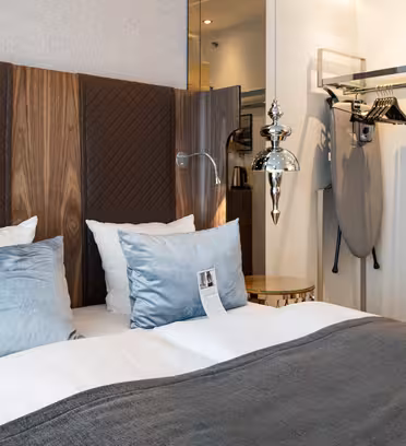 En pænt redt seng med gråt og hvidt sengetøj, flankeret af lyseblå puder, er på et moderne hotelværelse. Værelset har en hovedgærde i træ, hængende lampe, strygebræt og et åbent skab med bøjler og en hylde.