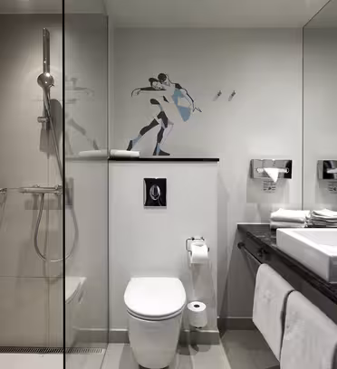 Moderne badeværelse med brusekabine i glas til venstre med regnbruser. Et toilet er i midten med toiletpapir. Til højre rummer en mørk bordplade en hvid vask og håndklæder. Et vægmaleri af en skater pryder væggen over toilettet.