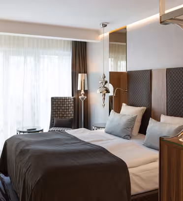 Et moderne hotelværelse med en stor seng klædt i hvidt og brunt sengetøj. Den har en mønstret lænestol, skrivebord med stol og dekorative lamper. Gulv til loft vinduer har gennemsigtige gardiner, og et lille bord står nær lænestolen.