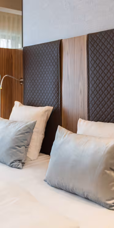 Et luksuriøst hotelværelse har en dobbeltseng med hvidt sengetøj, grå dekorative puder og en mørk quiltet sengegavl. Moderne sølvlamper hænger på hver side, og der er en mønstret stol og træborde i nærheden. Blødt lys filtrerer gennem gardiner.