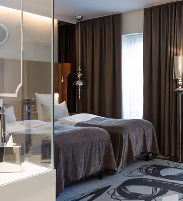 Hotelværelse med to pæne enkeltsenge, mørke gardiner og moderne design. Ved sengene står en håndvask i glas, og en mønstret lænestol er placeret tæt på væggen. Indretningen har neutrale toner og moderne belysning.