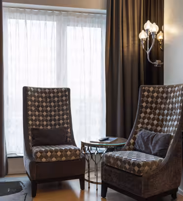 Et hyggeligt hotelværelse har to højryggede mønstrede stole omkring et lille glasbord. En moderne lampe står i nærheden, og et vægmonteret tv er synligt. Gennemsigtige og mørke gardiner dækker et stort vindue, så der kommer naturligt lys ind i rummet.