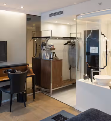 Moderne hotelværelse med vægmonteret tv, skrivebord og stol og et synligt badeværelse adskilt af en glasvæg. Det åbne klædeskab inkluderer en kaffemaskine og en hængende tøjtaske. Neutrale toner skaber en slank, minimalistisk æstetik.