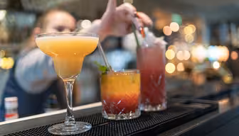 En bartender rækker ud efter at blande en pink cocktail med et sugerør. I forgrunden er der tre livlige cocktails på en bardisk: en gul margarita, en lagdelt orange og rød drink og den lyserøde cocktail. Baggrunden er blødt sløret.