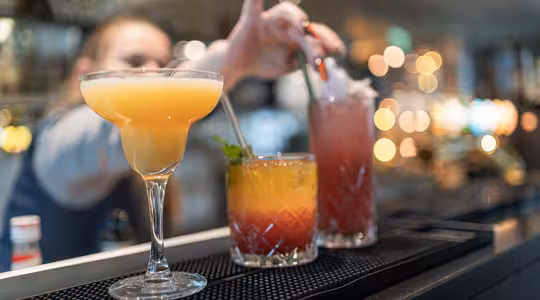 En bartender rækker ud efter at blande en pink cocktail med et sugerør. I forgrunden er der tre livlige cocktails på en bardisk: en gul margarita, en lagdelt orange og rød drink og den lyserøde cocktail. Baggrunden er blødt sløret.