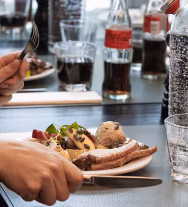 En person holder en gaffel og kniv over en tallerken med skiver kød, salat og en kartoffel. Flasker med cola og glas vand er synlige. Andre spisende gæster og tallerkener med mad er i baggrunden på et sort bord.