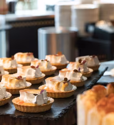 En udstilling af små syrlige kager toppet med hvirvler af hvid creme og små spiselige blomster, arrangeret på mørke serveringsbakker. I baggrunden er der stakke af hvide tallerkener og andre desserter.
