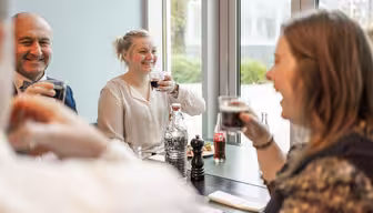 En gruppe mennesker sidder rundt om et bord ved et vindue, smiler og holder drinks. Der er vandflasker, en sodavandsflaske og en peberkværn på bordet. Baggrunden viser grønt udenfor.