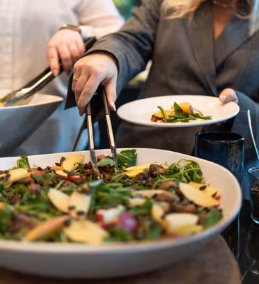To personer serverer sig selv ved en buffet med store skåle salat. En person bruger en tang til at plukke salat på en tallerken. Salaten toppes med æbleskiver og diverse grønt. En lille krukke med frø er i nærheden.