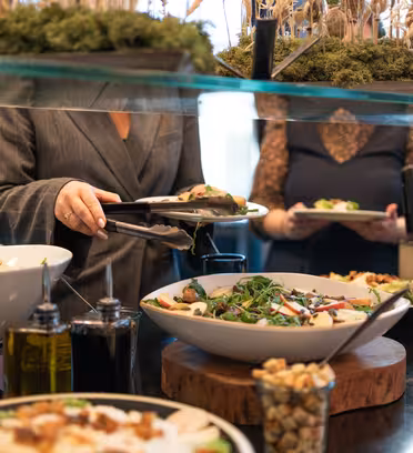 Folk serverer sig selv fra en buffet med skåle med salat, med forskellige toppings og dressinger. Rammerne er moderne og dekorative, med planter og olivenolieflasker synlige.