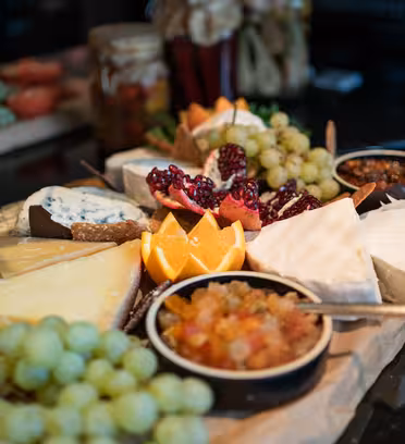 Et charcuteribræt med en række forskellige oste, vindruer, appelsinskiver, halve granatæble og små retter med chutney og syltetøj. Baggrunden viser et sløret billede af andre madvarer.