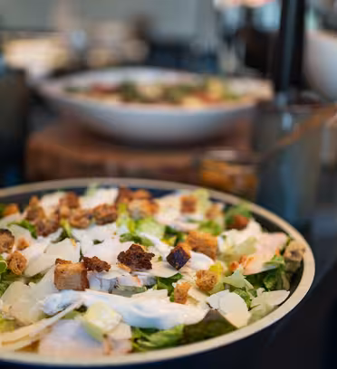Et nærbillede af en Cæsarsalat toppet med croutoner og barberet ost i en skål. Andre retter er slørede i baggrunden. Indstillingen ser ud til at være en restaurant eller buffet.