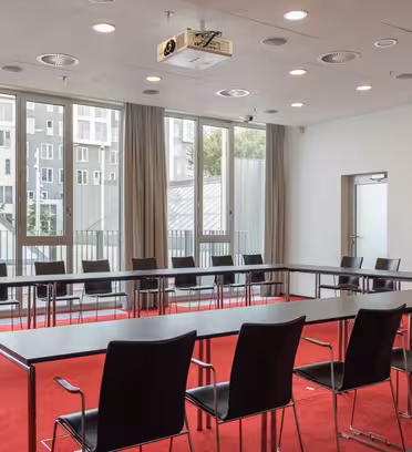 Moderne mødelokale med rød løber med U-formede sorte borde og stole. Store vinduer giver naturligt lys, med en projektor på loftet og udsigt til bygninger udenfor. Hvide vægge med minimal indretning.