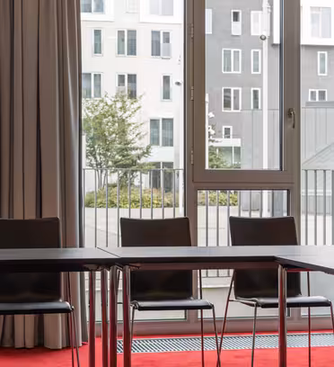 Mødelokale med et U-formet arrangement af borde og stole på en rød løber. Store vinduer med beige gardiner afslører udsigt til moderne hvide og grå bygninger.