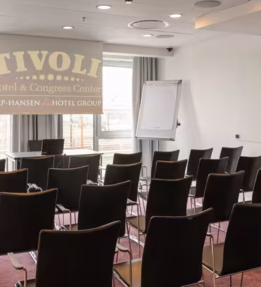 Et lille mødelokale med rækker af tomme sorte stole ud mod en projektorskærm med "Tivoli Hotel & Congress Center". Der er en flipover og et stort vindue med hvide gardiner, der lukker naturligt lys ind.