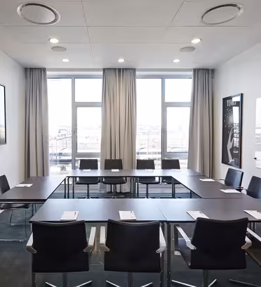 Et moderne mødelokale med U-formet bordopstilling, sorte stole, et stort væghængt tv, whiteboard og høje vinduer med gardiner. Notesblokke og kuglepenne er på bordet, og bybilledet er synligt uden for vinduerne.