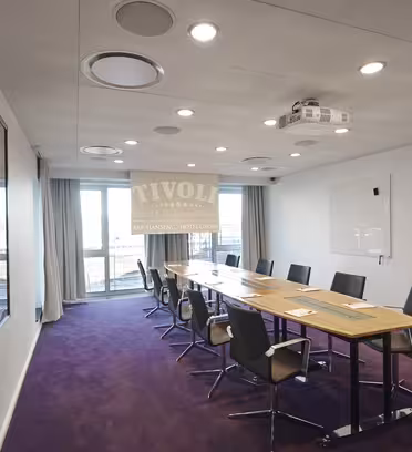 Et moderne mødelokale med et langt træbord omgivet af ni sorte stole. Rummet har en whiteboardtavle, en loftmonteret projektor og store vinduer med udsigt til et bylandskab. Lilla tæppe dækker gulvet.