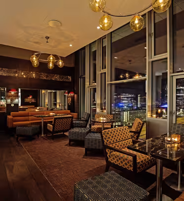 En moderne, svagt oplyst lounge med gulv til loft-vinduer, der viser et bybillede om natten. Rummet har læder og mønstrede siddepladser, mørke borde, pendellamper og små levende lys på borde, hvilket skaber en hyggelig, eksklusiv atmosfære.