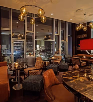 En stilfuld lounge med lædersofaer og moderne belysning. Marmorborde er arrangeret med komfortable stole. Store gulv-til-loft-vinduer tilbyder udsigt over bybilledet. Varm belysning skaber en indbydende atmosfære.