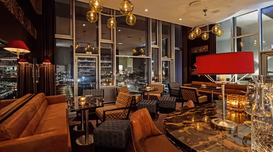 En stilfuld lounge med lædersofaer og moderne belysning. Marmorborde er arrangeret med komfortable stole. Store gulv-til-loft-vinduer tilbyder udsigt over bybilledet. Varm belysning skaber en indbydende atmosfære.