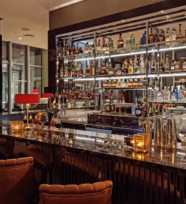 En luksuriøs bar med marmorbordplade, røde lamper og plysbrune stole. De bagerste hylder viser forskellige flasker spiritus, oplyst af blød, omgivende belysning.
