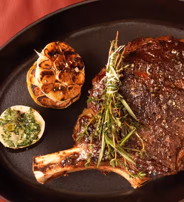 En kogt tomahawk bøf pyntet med urter sidder på et sort ovalt fad. Ved siden af er en ristet hvidløgsløg og en halv citron med urtesmør. Fadet placeres på en rød læderoverflade.