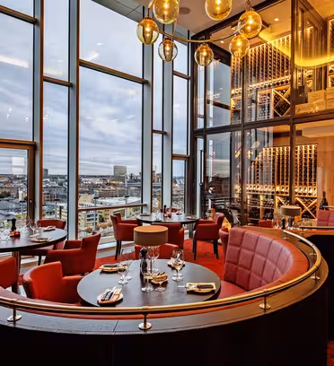 Luksuriøs restaurant med høje vinduer med udsigt over byen. Med røde polstrede kabiner, runde borde til spisning, varm belysning og en dekorativ vinudstillingsvæg. Plys røde tæpper komplementerer den elegante stemning.