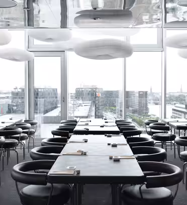 En moderne restaurant med sorte borde og stole vises. Elegante hvide lysarmaturer hænger fra loftet. Store vinduer giver udsigt over et bybillede med bygninger og en vandmasse synlig i baggrunden.