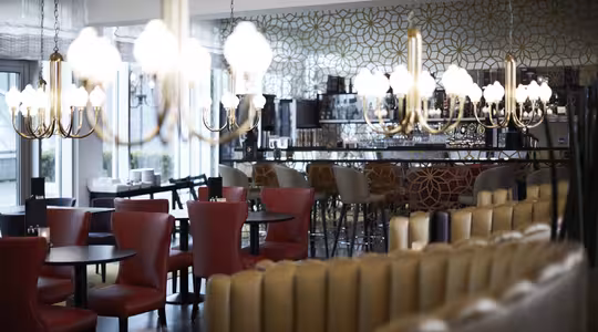 Et moderne restaurantinteriør med røde og beige stole, runde borde og et barområde. Elegante lysekroner hænger fra loftet med en geometrisk mønstret væg i baggrunden. Atmosfæren er chik og sofistikeret.