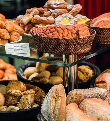 Diverse kager og brød vist på en glashylde, inklusive croissanter, kager i pulverform og forskellige brød. De er arrangeret i flettede kurve og runde bakker, hvilket skaber en tiltalende og mangfoldig bagerpræsentation.