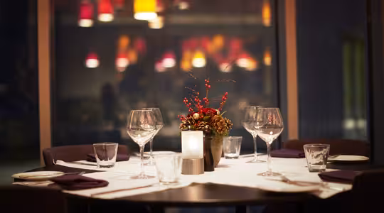 Et svagt oplyst restaurantbord er elegant dækket med fire vinglas, vandglas og tallerkener omkring et midtpunkt af røde bær og stearinlys. Varme, farverige pendellamper hænger i baggrunden og skaber en hyggelig atmosfære.