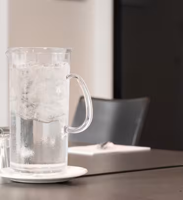 En glaskande fyldt med isvand sidder på en underkop oven på et mørkt bord. Flere op-ned-ned-glas er i nærheden. Sorte stole og en delvist synlig dør er i baggrunden.