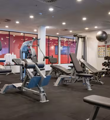 Et moderne fitnesscenter med forskellige træningsredskaber, herunder vægtmaskiner, bænke og et stativ med håndvægte. Rummet har røde detaljer, højt til loftet med lys og store vinduer med matteret glas.