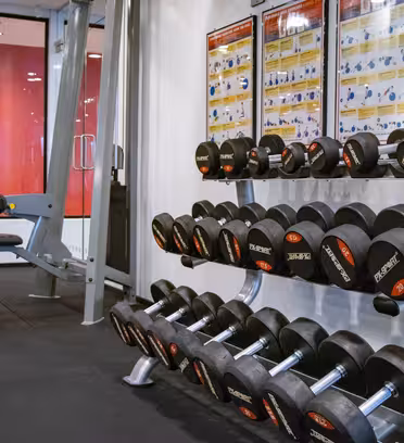 Et fitnesscenterinteriør med et stativ med diverse håndvægte og en vægtløftningsbænk til venstre. Træningsinstruktionsplakater hænger på væggen over udstyret. Rummet har sort gulv og røde accenter i baggrunden.