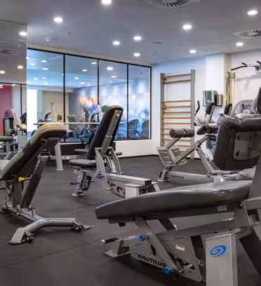 Et moderne fitnesscenter interiør med forskellige træningsmaskiner, herunder en siddende bænk og benpres. Væggene har spejle og vinduer. Rummet er godt oplyst med ovenlys, og gulvet er belagt med sorte gummimåtter.