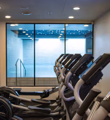 Række af elliptiske maskiner i et fitnesscenter med front mod et vægmonteret tv. Bag en glasparti er der udsigt til et tilstødende swimmingpoolområde. Rummet er godt oplyst og har et moderne design.