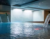 Indendørs swimmingpool med to vægmonterede vandelementer og en buet vandfaldstud. Væggene er beklædt med lyse fliser, og vandet har en rolig, reflekterende overflade. Loftslamper giver en blød stemning.