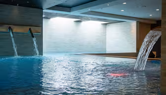 Indendørs swimmingpool med to vægmonterede vandelementer og en buet vandfaldstud. Væggene er beklædt med lyse fliser, og vandet har en rolig, reflekterende overflade. Loftslamper giver en blød stemning.