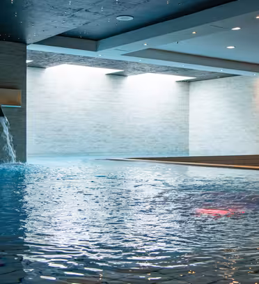 Indendørs swimmingpool med to vægmonterede vandelementer og en buet vandfaldstud. Væggene er beklædt med lyse fliser, og vandet har en rolig, reflekterende overflade. Loftslamper giver en blød stemning.