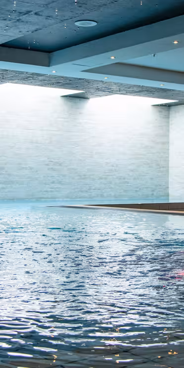 Indendørs swimmingpool med to vægmonterede vandelementer og en buet vandfaldstud. Væggene er beklædt med lyse fliser, og vandet har en rolig, reflekterende overflade. Loftslamper giver en blød stemning.