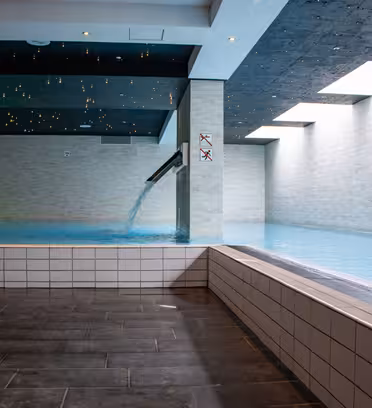 Indendørs pool med brusende vandfald og lyse stenvægge. Loftet har små stjernelignende lys, der skaber en stjerneklar effekt. Sollys strømmer gennem en række ovenlys langs den ene side og oplyser vandet.