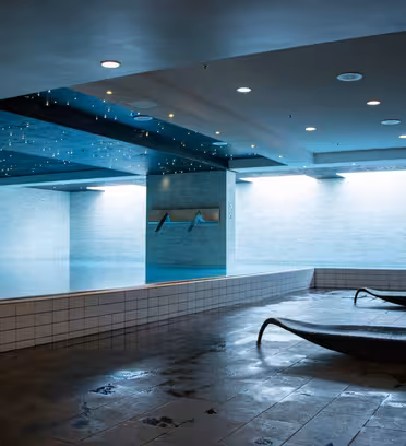 Indendørs swimmingpool med moderne design. Poolområdet har skarp belysning, klinkegulv og to liggestole. Et loft med stjernelignende lys tilføjer et dekorativt touch, og advarselsskilte er synlige på en væg.