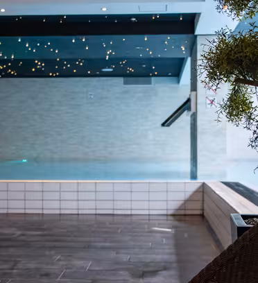 Indendørs swimmingpoolområde med dæmpet belysning, med en stjerneklar loftseffekt. Der er et lille træ til højre og sort klinkegulv. Poolen har en klar, rolig overflade, der skaber en rolig atmosfære.