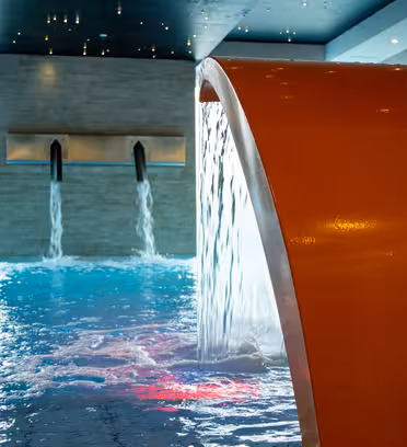 Indendørs pool med brusende vand. En stor orange struktur buer over poolen og frigiver vand ved siden af to springvand på den fjerneste væg. Indstillingen har en moderne og fredfyldt atmosfære med blid belysning, der reflekterer fra vandet.