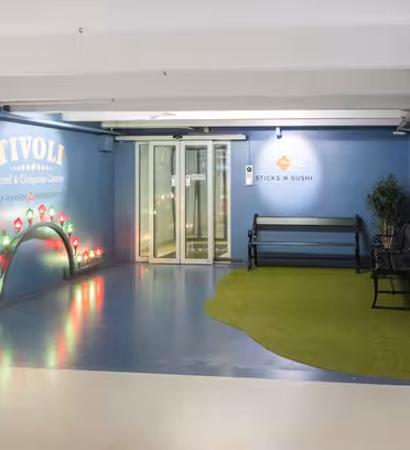 Indgangsparti med en blå væg med skiltning fra Tivoli Hotel & Congress Center. Der er en lille bro med rødt lys og en bænk på en plet af kunstgræs. Sticks'n'Sushi-skiltet er synligt ved siden af en glasdør.